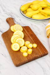 Sliced Yellow Crookneck Squash on a Wood cutting Board on Marble Countertop with a White Bowl of Uncut Squash in Background