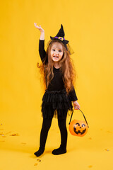 little girl with long red hair smiles in a Halloween witch costume and holds a pumpkin-shaped candy...
