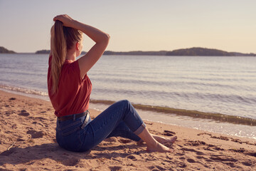 Fototapeta na wymiar Beautiful young white woman is sitting on the sandy beach in sunlight in blue jeans and red shirt and looking at the sunset. Copy space.