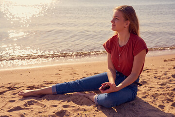 Fototapeta na wymiar Happy and beautiful young white woman is sitting on the sandy beach in sunlight in blue jeans and red shirt. Copy space.