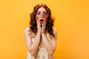 Close-up portrait of red-haired girl with wild flowers in her hair looking surprised into camera. Woman in lilac glasses posing on isolated background