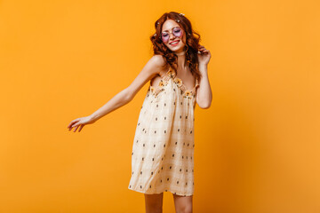 Portrait of charming girl in short dress and lilac glasses. Woman with wildflowers in her hair dancing on orange background