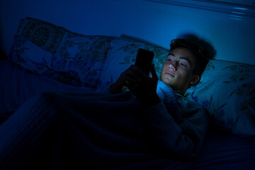 one addicted teenager at the phone using it late night in his bedroom under the blanket in his bed - social media and network addicted concept and lifestyle