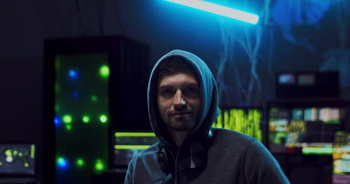 Close up of the young attractive Caucasian guy hacker in a hood looking straight to the camera and smiling cheerfully while sitting in the dark server room with computers on the digital world