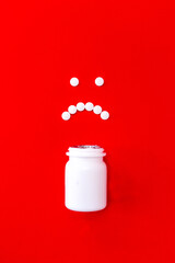 Unhappy face made of white circular pills with a white pill plastic bottle on a red background