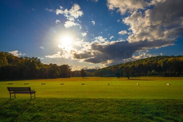 This idyllic image shows the sun setting over an empty golf course with an inviting empty bench and beautiful clouds in a the blue sky.