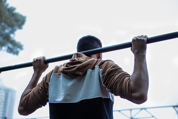 Naklejka premium Young man pulling up, warming up on street workout area. Physical activity outdoors, caucasian man with tanned skin training, cloudy cold day, sport casual clothes