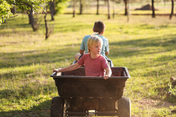 Young boy riding in trailer of ride on lawn mower being driven by his big brother on country...