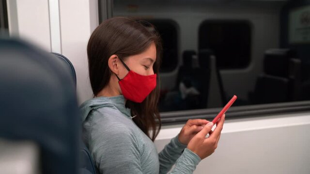 Woman wearing face mask in public transportation during coronavirus Covid-19 pandemic. Train transport commuter concept. Multiracial woman passenger using smart phone with face covering on commute