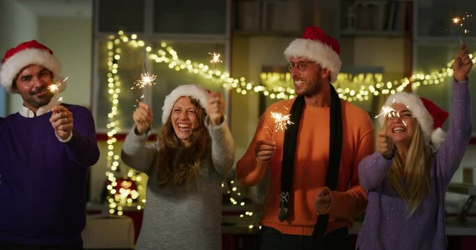 Authentic shot of happy cheerful friends wearing santa hats singing a Christmas Song with sparklers in their hands while celebration. Concept of family, friendship, winter holidays,Christmas, present