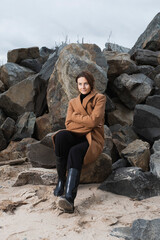 Portrait of a beautiful young model in warm clothes enjoy day, on background rocks and sea  in  sunny autumn day . Autumn warm photo. Woman smiling and look away, joyful cheerful mood.