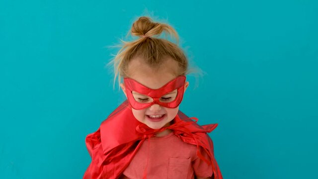 Funny blonde toddler girl in red superhero costume with mask looks smiling and waves hand against blue studio wall closeup