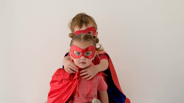 Funny boy in coloured superhero costume hugs and kisses little sister in red mask sitting against white background closeup