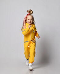 Sporty blonde kid baby girl with colorful braids wearing stylish yellow jumpsuit and sneakers runs towards camera