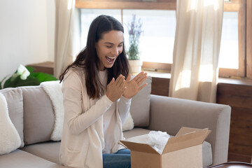 Happy laughing girl teenager sitting on couch clapping hands in delight opening cardboard box with birthday present delivered by courier, young female client getting purchase from online beauty store