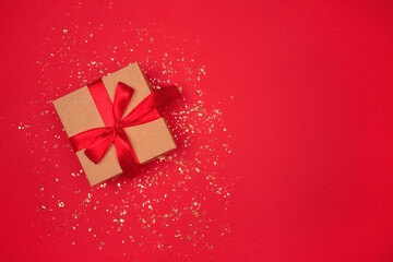 Present box with red bow on bright red background with golden sparkles around and copyspace for your text. Festive backdrop for your project. Flat lay style. Christmas or birthday celebration concept