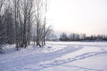 Winter landscape - white snow with traces of shoes and skis on the field. The ski track and road skirting the forest with bare trees, soft sunlight.