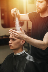 Caring hairdresser applying spray to young man's hair in a barber shop