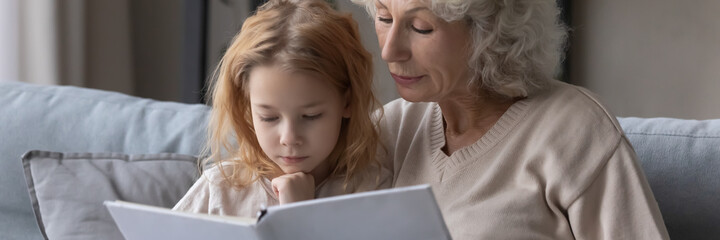 Wide image close up mature grandmother and little granddaughter reading book together, caring loving granny embracing preschool girl child, telling interesting fairytale story, children education - Powered by Adobe