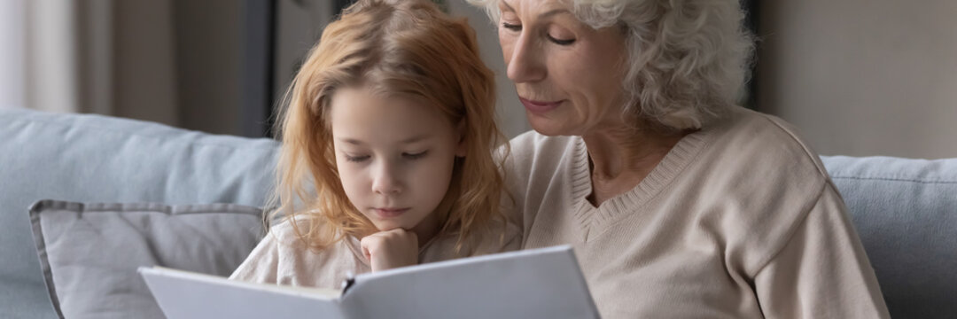 Wide image close up mature grandmother and little granddaughter reading book together, caring loving granny embracing preschool girl child, telling interesting fairytale story, children education