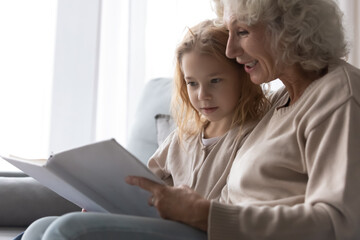 Close up smiling mature grandmother reading book to little granddaughter, telling interesting fairytale story, loving elderly woman hugging pretty preschool girl, relaxing on cozy couch at home