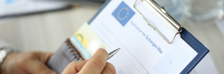 Close-up of person filling application form for schengen visa on clipboard. Man holding silver pen....