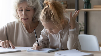 Close up mature grandmother helping unhappy little granddaughter with school homework, difficult tasks, elderly tutor teacher teaching pupil, sitting at desk together, angry girl studying at home