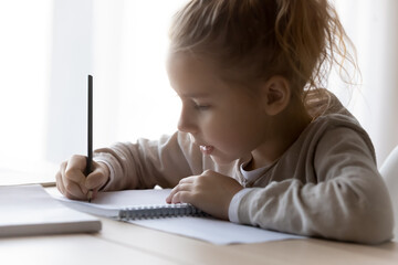 Close up serious little girl writing in notebook, holding pencil, studying at home, sitting at desk, focused busy child schoolgirl preparing school homework, assignments, homeschooling concept