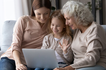 Fototapeta na wymiar Happy little girl with mother and grandmother using laptop together, sitting on couch, looking at screen, family making video call, smiling pretty child waving hand, saying hello, greeting relatives