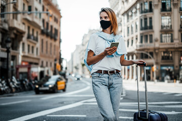 Young traveling woman with suitcase on a sunny city street. Traveler wearing mask on vacation.