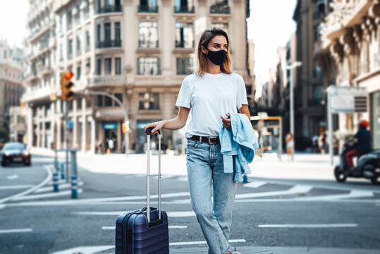 Young traveling woman with suitcase on a sunny city street. Traveler wearing mask on vacation.