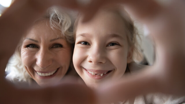 Close up happy mature grandmother and granddaughter showing heart gesture, looking at camera through fingers, smiling elderly woman and little girl having fun, charity and love concept