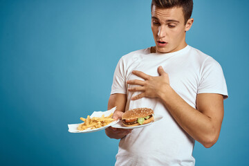 young guy with fries and hamburger on blue background interested look emotions fast food calories cropped view Copy Space