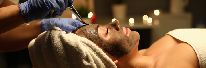 Close-up of peaceful and calm young woman getting skin treatment with clay mask. Female covered with white towel. Joy and relax at cosmetologist. Beauty and spa day concept