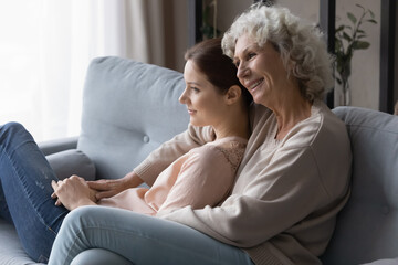 Smiling mature woman with grownup daughter enjoying leisure time at home, hugging, relaxing on cozy couch in living room, happy grandmother and granddaughter dreaming, visualizing together