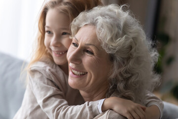 Close up smiling pretty little girl hugging mature grandmother, family enjoying tender moment, sitting on cozy couch at home, overjoyed loving elderly grandma and preschool girl cuddling