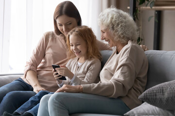 Fototapeta premium Happy little girl with mother and grandmother using smartphone together, looking at screen, watching funny video in social network, chatting or shopping online, family enjoying leisure time at home