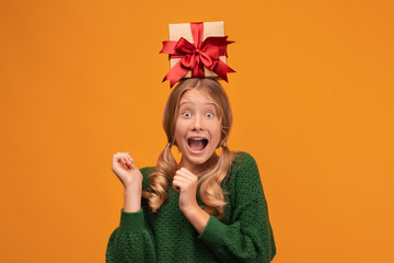 Charming blonde teen hold gift box on her head. Studio shot, yellow background. New Year Birthday Holiday concept