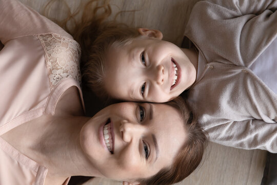 Top view head shot portrait smiling mother and little daughter lying on warm floor, looking at camera, pretty girl child and happy young mum posing for funny family photo at home, having fun together