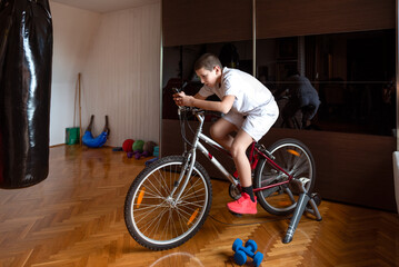 Young boy training on bicycle stationery trainer in improvised home gym during quarantine holding mobile phone and watching online training; spinning class