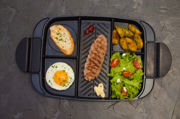 Breakfast, fried steak or cutlet, salad with tomato and green vegetables, toast, potatoes, egg on a black plate. Concept of a healthy diet, diet, menu, good nutrition.