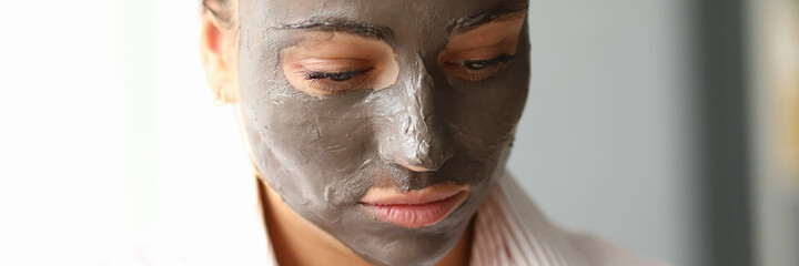 Fototapeta premium Close-up of young pretty female with clay mask on face. Attractive woman in pyjamas after night sleep. Copy space in left side. Morning facial treatment and relax concept