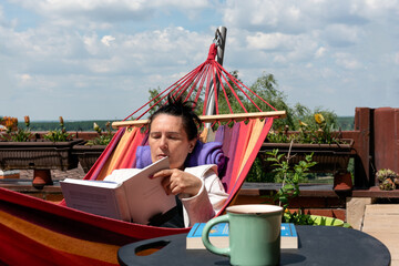 Woman lying in colorful hammock on terrace, reading book, enjoying her free time and nice weather