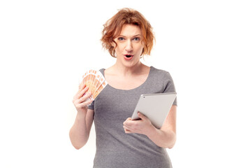 Red haired adult mature woman in gray dress with a tablet and money in her hands, very enthusiastic