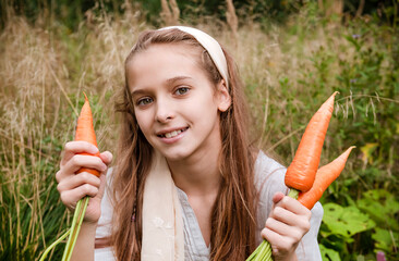 girl holds fresh carrots in her hands against a background of greenery