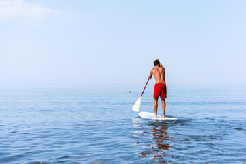 Athletic young man paddling on a sup board on the quiet sea - Stand up paddle boarder training on a rowing board on a flat calm sea - Back view and copy space for text