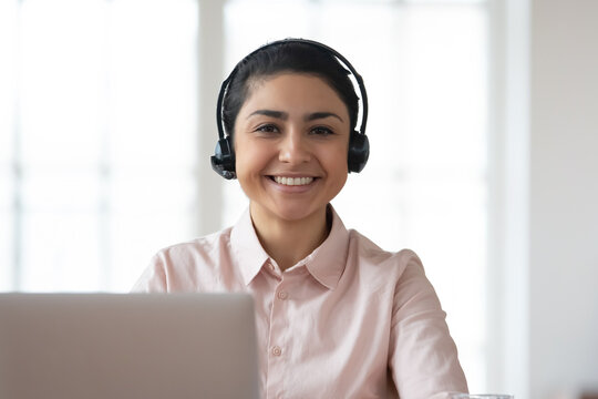 Headshot portrait of smiling young indian female employee in headphones have webcam conference on laptop in office. Profile picture of happy millennial woman in earphones talk speak on video call.