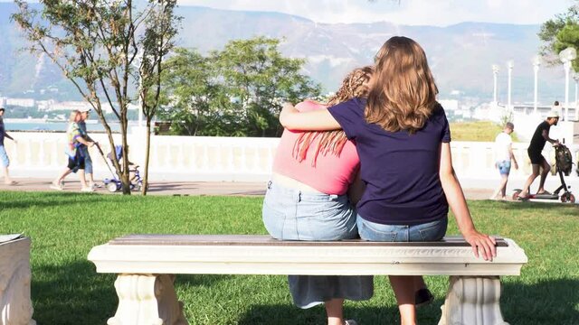 Rear view of couple of girlfriends sitting on a bench relaxing under the sun near the sea coast. Media. Two women lesbians sitting and hugging near green lawn on a sunny summer day, LGBT concept.