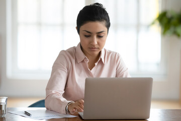 Serious millennial indian female employee sit at desk in office typing texting on laptop. Focused young ethnic woman worker busy using computer, consult business client online. Communication concept.