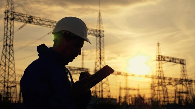 An electrical engineer wearing a helmet works with a tablet near high voltage electrical lines. An electrician works with the documents of an electrical substation. Energy business concept.